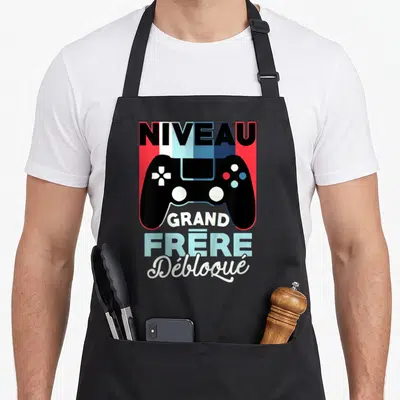 Tablier de cuisine noir avec illustration manette et texte Niveau Grand Frère Débloqué en couleurs vives sur fond géométrique.