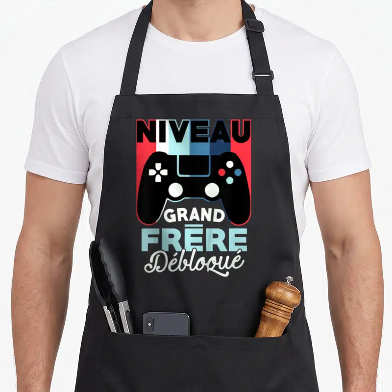 Tablier de cuisine noir avec illustration manette et texte Niveau Grand Frère Débloqué en couleurs vives sur fond géométrique.