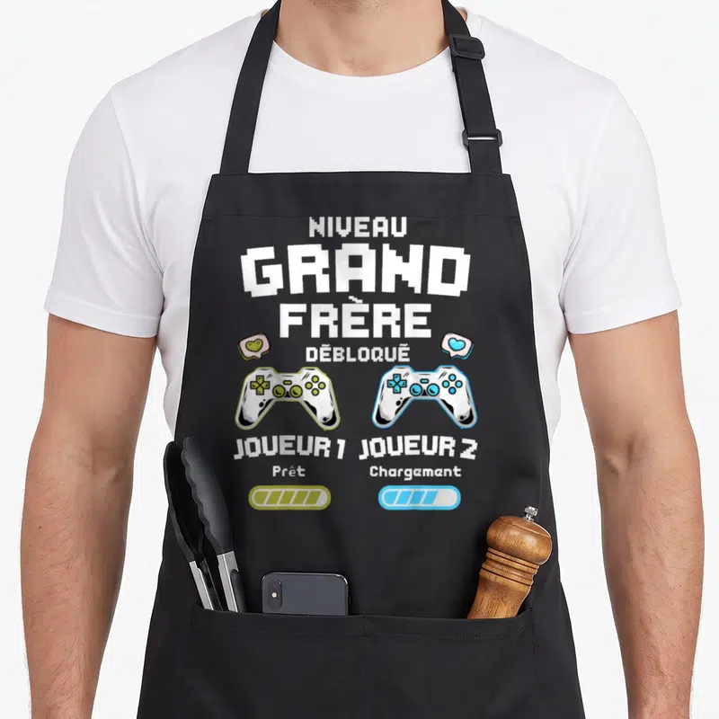 Tablier de cuisine noir avec motif Niveau Grand Frère Débloqué, deux manettes de jeu vidéo et barres de progression colorées.