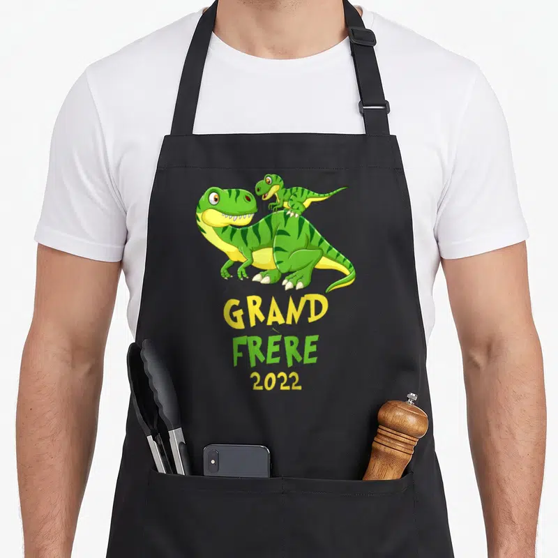 Tablier de cuisine noir avec deux dinosaures verts et le texte Grand Frère 2022 imprimé en jaune et vert.