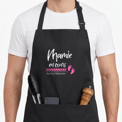 Tablier de cuisine noir avec inscription Mamie en cours Veuillez Patienter et deux petits pieds roses stylisés.
