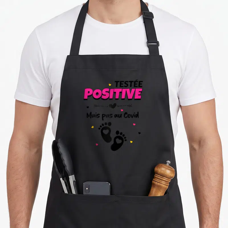 Tablier de cuisine noir avec l'inscription Testée Positive Mais pas au Covid et des empreintes de pieds roses, illustrant un motif humoristique sur la grossesse.