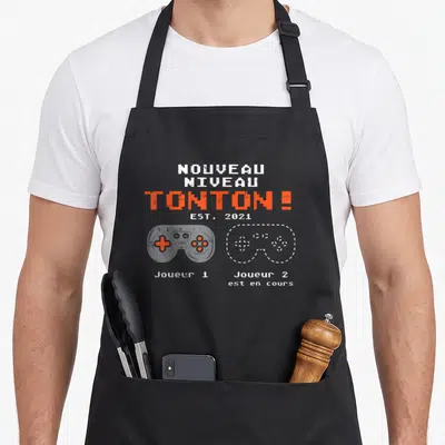 Tablier de cuisine noir avec inscription Nouveau Niveau Tonton Est 2021 et deux manettes de jeu vidéo stylisées en pixel art.