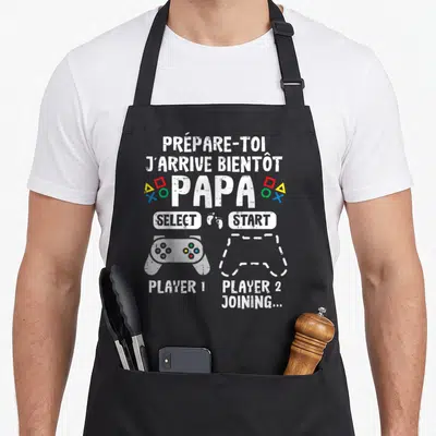 Tablier de cuisine noir avec l'inscription Prépare-toi j'arrive bientôt papa et une illustration de manette de jeu vidéo Player 2 joining.
