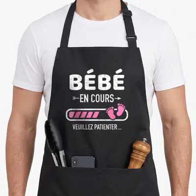 Tablier de cuisine noir avec l'inscription Bébé en cours et deux petits pieds roses sur une barre de chargement.