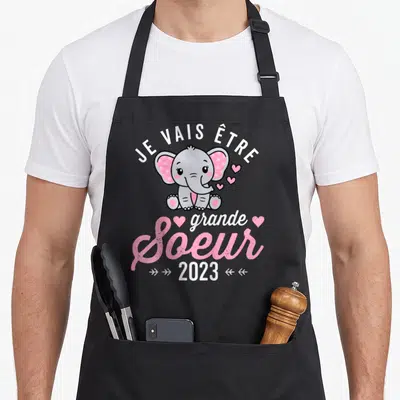 Tablier de cuisine noir avec un éléphant rose et le texte Je vais être grande soeur 2023 imprimé en blanc et rose.