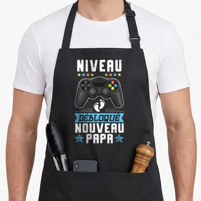 Tablier de cuisine noir avec illustration manette de jeu et texte Niveau Nouveau Papa Débloqué en blanc et bleu.