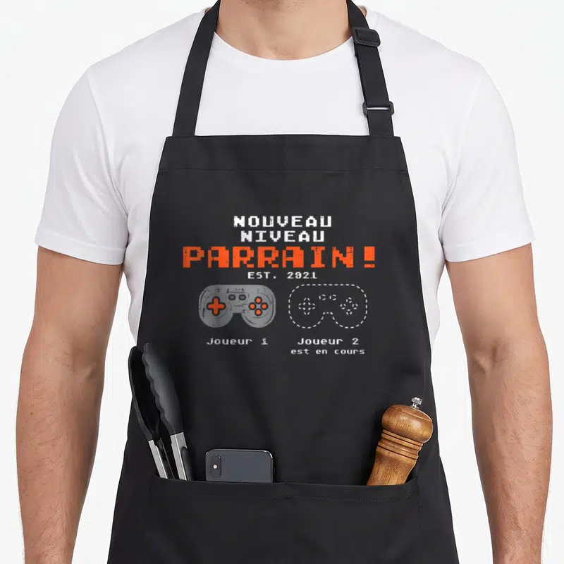 Tablier de cuisine noir avec inscription Nouveau Niveau Parrain Est 2021 et deux manettes de jeu vidéo stylisées en orange et blanc.
