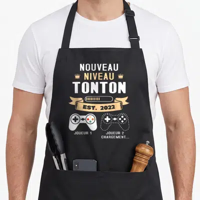 Tablier de cuisine noir avec inscription Nouveau Niveau Tonton Est 2022 et deux manettes de jeu vidéo Joueur 1 et Joueur 2 Chargement.