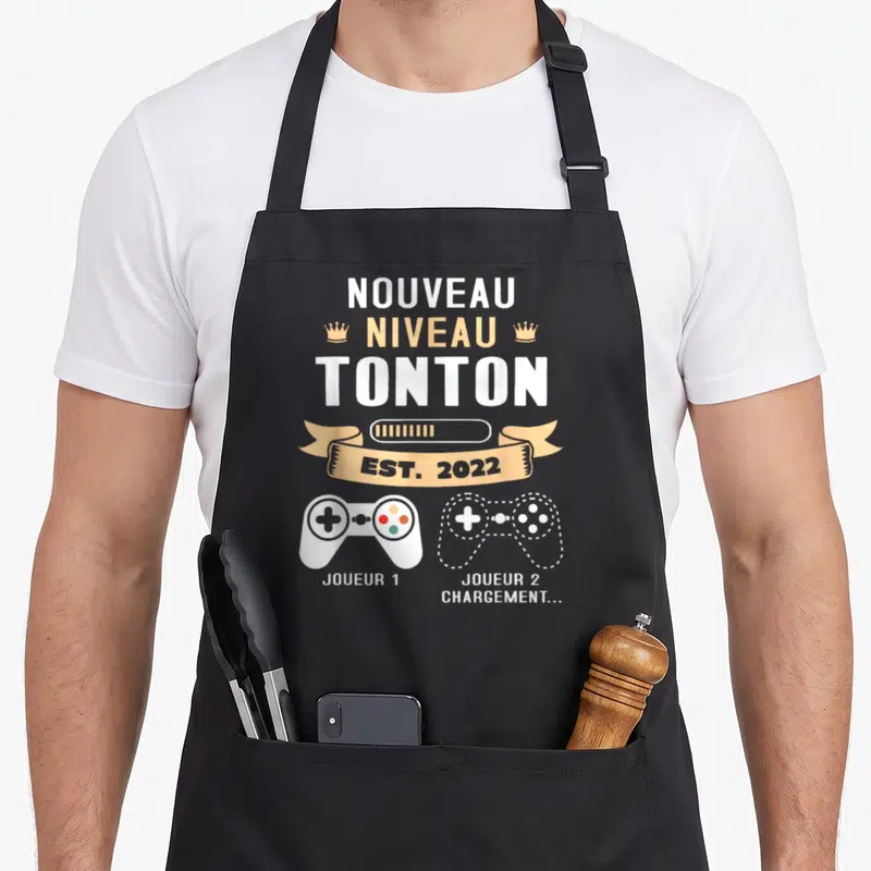 Tablier de cuisine noir avec inscription Nouveau Niveau Tonton Est 2022 et deux manettes de jeu vidéo Joueur 1 et Joueur 2 Chargement.