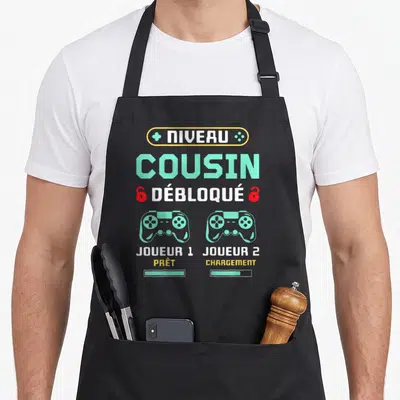 Tablier de cuisine noir avec inscription Niveau Cousin Débloqué et deux manettes de jeu vertes.