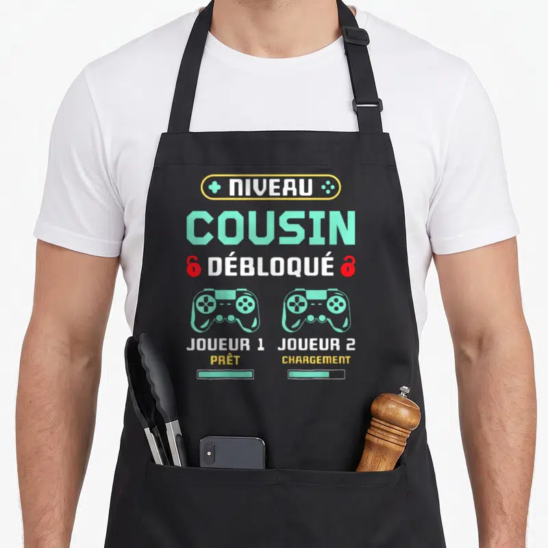 Tablier de cuisine noir avec inscription Niveau Cousin Débloqué et deux manettes de jeu vertes.