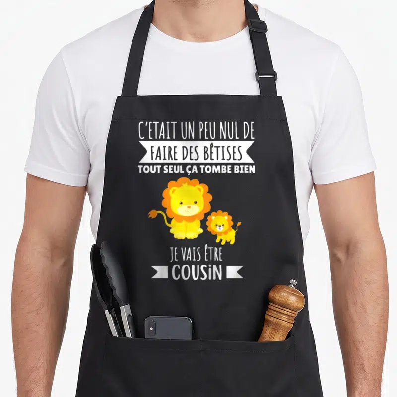 Tablier de cuisine noir avec deux lions jaunes et le texte Je vais être cousin 2027 imprimé en blanc.