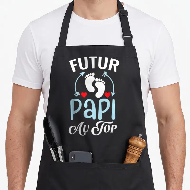 Tablier de cuisine noir avec l'inscription Futur Papi Au Top et des petits pieds blancs entourés de flèches bleues et de cœurs rouges.