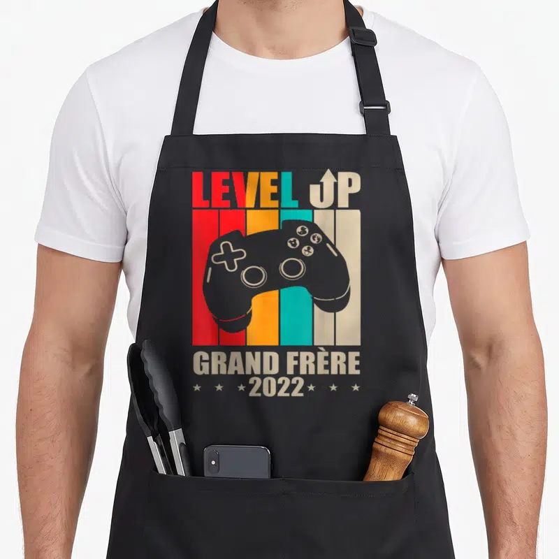 Tablier de cuisine noir avec manette de jeu et texte Level Up Grand Frère 2022 sur fond de bandes colorées.