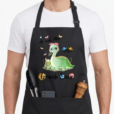 Tablier de cuisine noir avec dinosaure vert, œuf éclos et texte Je vais être Grande Soeur 2022 entouré de papillons colorés.