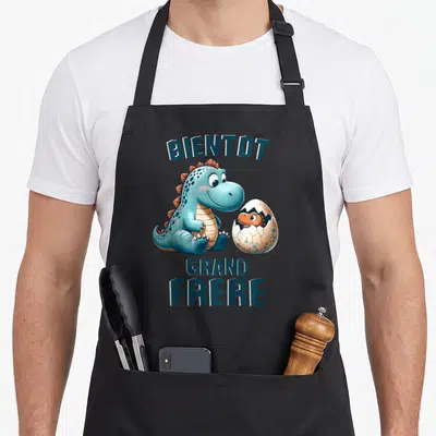 Tablier de cuisine noir avec illustration dinosaure bleu tenant un œuf et texte Bientôt Grand Frère.