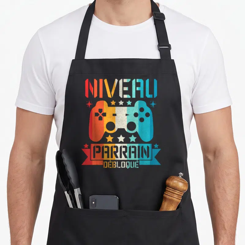 Tablier de cuisine noir avec illustration manette de jeu vidéo multicolore et texte Niveau Parrain Débloqué en typographie rétro.