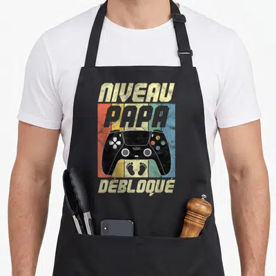 Tablier de cuisine noir avec manette de jeu et inscription Niveau Papa Débloqué sur fond coloré.