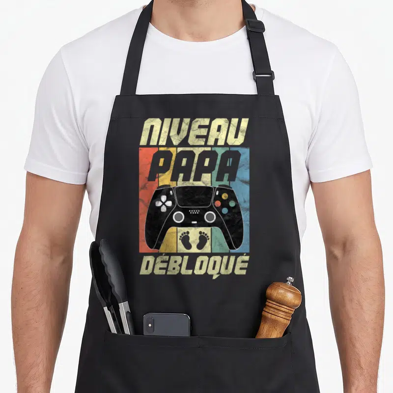 Tablier de cuisine noir avec manette de jeu et inscription Niveau Papa Débloqué sur fond coloré.