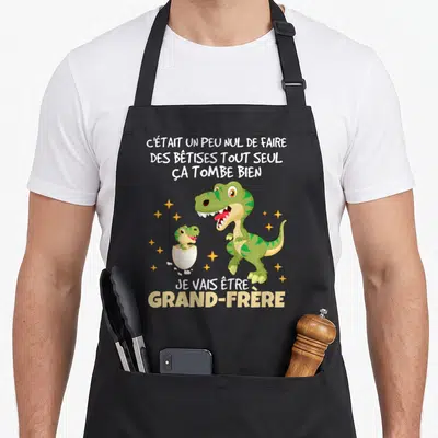 Tablier de cuisine noir avec deux dinosaures verts et le texte annonce naissance futur grand frère 2026