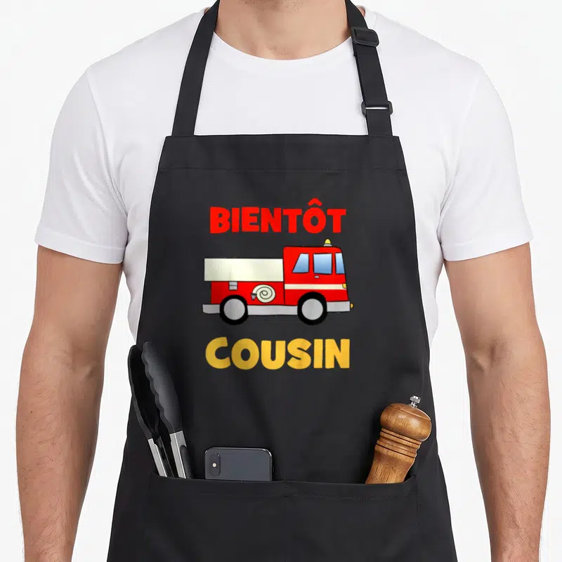 Tablier de cuisine noir avec un camion de pompier rouge et les mots Bientôt Cousin en lettres colorées.