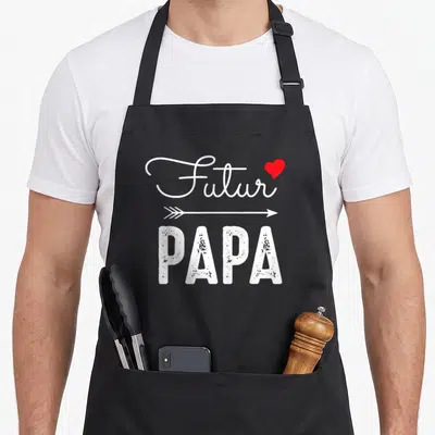 Tablier de cuisine noir avec l'inscription Futur Papa en typographie blanche et une flèche horizontale souscrite par un petit cœur rouge vif.