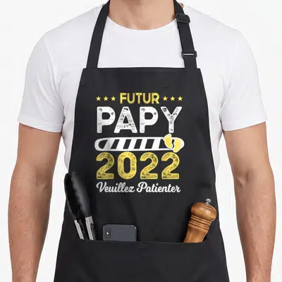 Tablier de cuisine noir avec inscription Futur Papy 2022 Veuillez Patienter et empreintes de pieds jaunes.