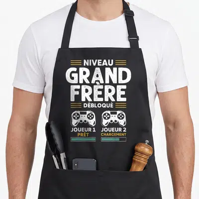 Tablier de cuisine noir avec texte Niveau Grand Frère Débloqué et deux manettes de jeu vidéo blanches et dorées.