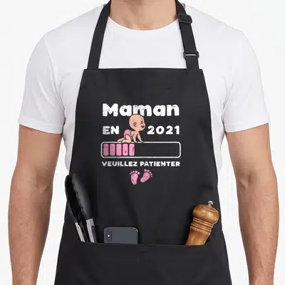 Tablier de cuisine noir avec illustration d'un bébé et texte Maman en 2021 Veuillez patienter en blanc et rose.