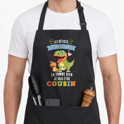 Tablier de cuisine noir avec deux dinosaures colorés et le texte Ça tombe bien je vais être cousin imprimé en lettres vives.