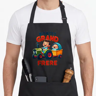 Tablier de cuisine noir avec un garçon au volant d'un tracteur vert tirant une remorque avec un bébé souriant.