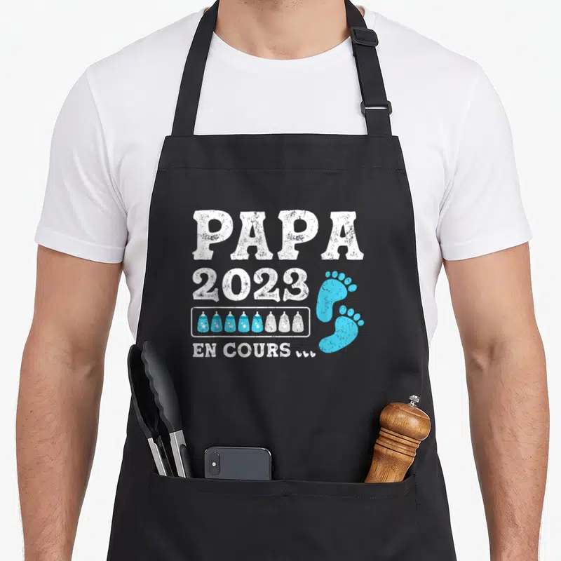 Tablier de cuisine noir avec inscription Papa 2023 En Cours et motifs de biberons bleus et blancs.
