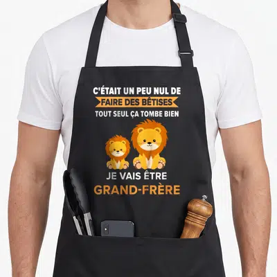 Tablier de cuisine noir avec deux lions et le texte Je vais être grand-frère en orange et blanc.