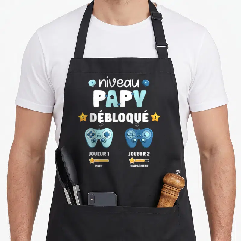 Tablier de cuisine noir avec deux manettes de jeu bleues et le texte Niveau Papy Débloqué Joueur 1 et 2.
