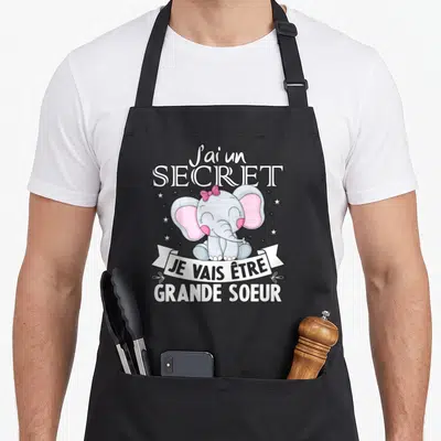 Tablier de cuisine noir avec un éléphant rose portant un nœud et le texte J'ai un secret je vais être grande soeur.