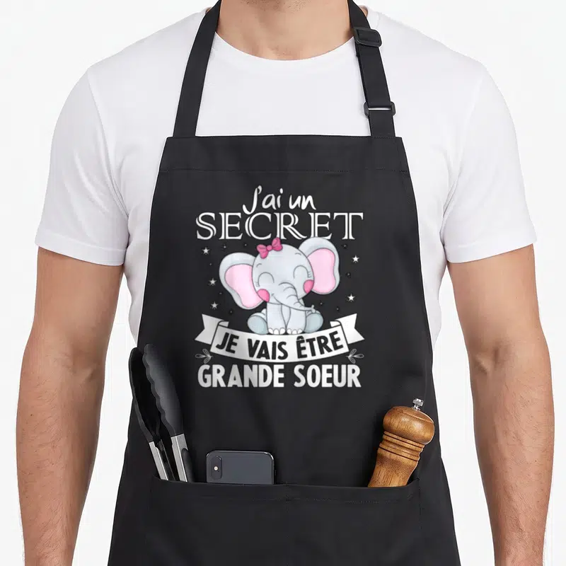 Tablier de cuisine noir avec un éléphant rose portant un nœud et le texte J'ai un secret je vais être grande soeur.