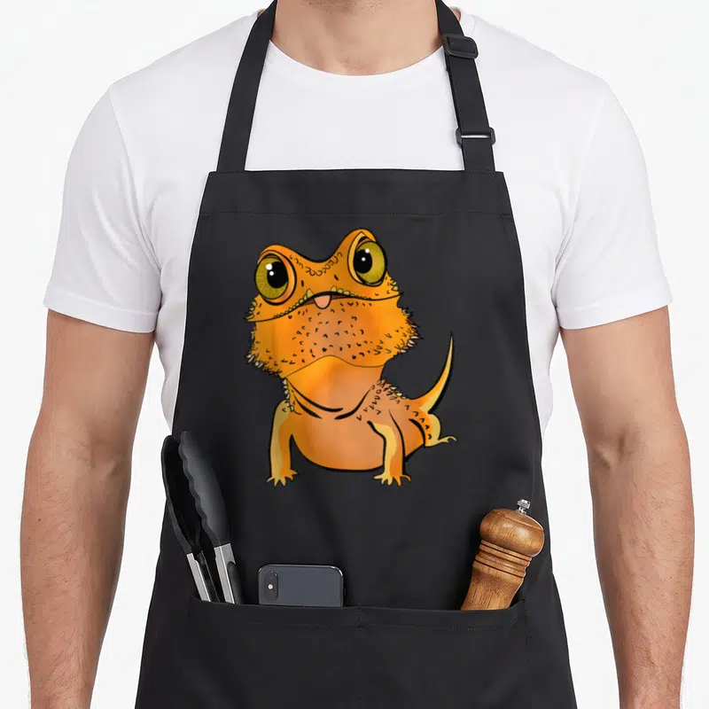 Tablier de cuisine noir avec illustration d'un bébé dragon barbu orange aux grands yeux jaunes et langue rose saillante.