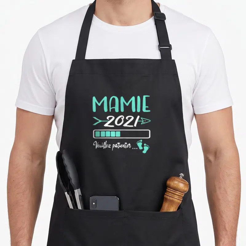 Tablier de cuisine noir avec inscription Mamie 2021, barre de chargement turquoise et petits pieds imprimés en bas à droite.