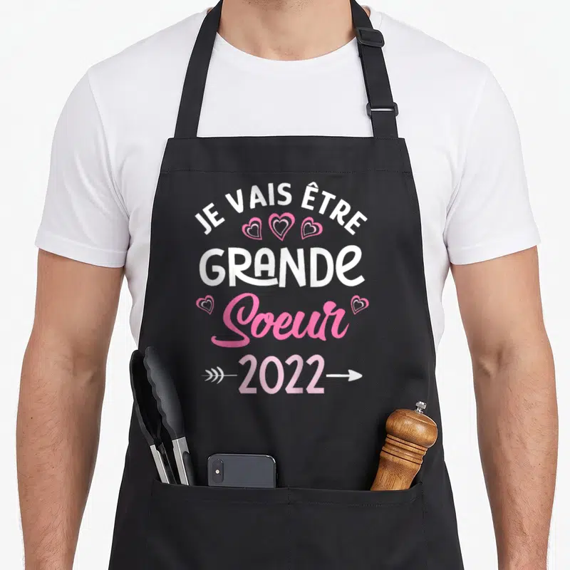 Tablier de cuisine noir avec inscription Je vais être Grande Soeur 2022 ornée de cœurs roses et de flèches directionnelles.