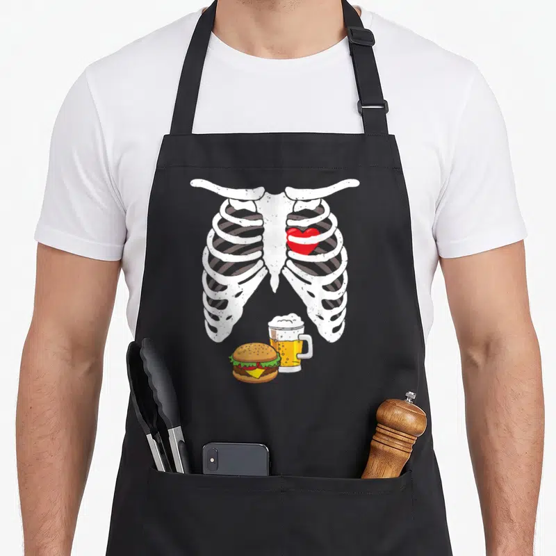 Tablier de cuisine noir avec illustration squelette, cœur rouge, burger et bière sur fond uni.