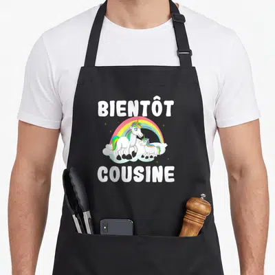 Tablier de cuisine noir avec inscription Bientôt Cousine et illustration de deux licornes sous un arc-en-ciel.