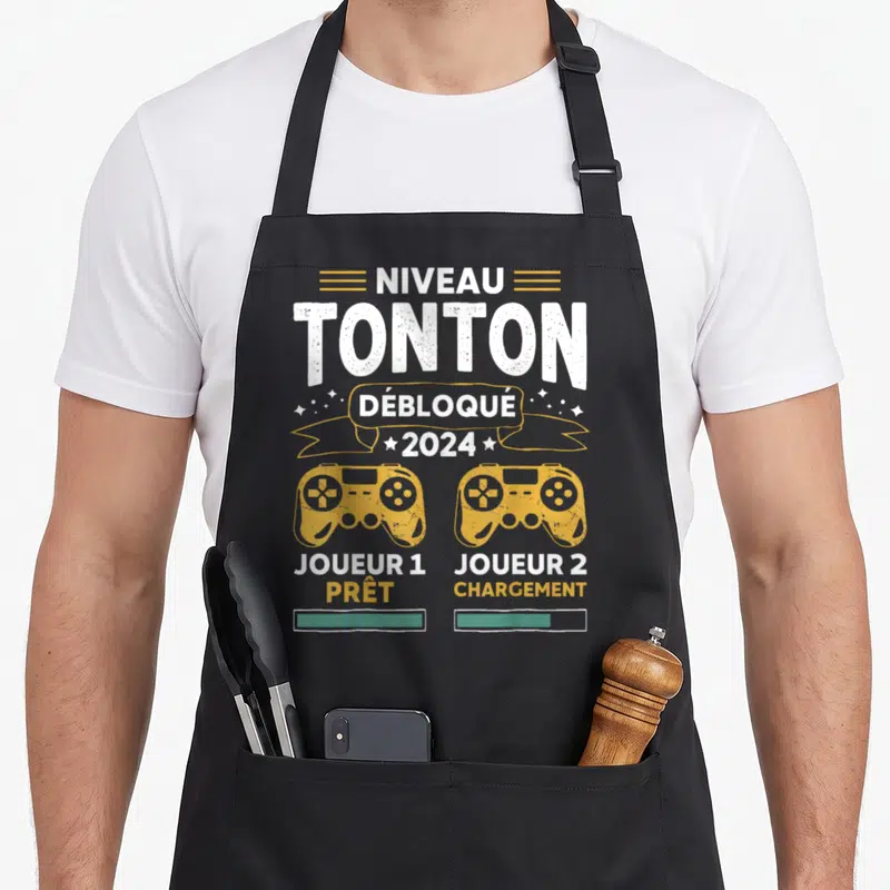 Tablier de cuisine noir avec inscription Niveau Tonton Débloqué 2024 et deux manettes de jeu vidéo jaunes.