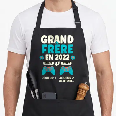 Tablier de cuisine noir avec l'inscription Grand Frère en 2022 et deux manettes de jeu vidéo bleues sur le devant.