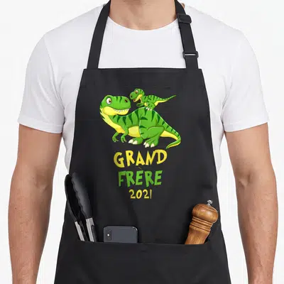 Tablier de cuisine noir avec deux dinosaures verts et jaunes et le texte Grand Frère 2021 imprimé au centre.