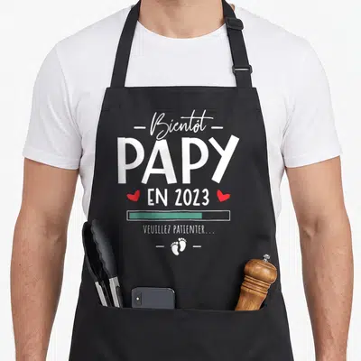 Tablier de cuisine noir avec texte blanc Bientôt Papy en 2023 et icônes de petits pieds rouges