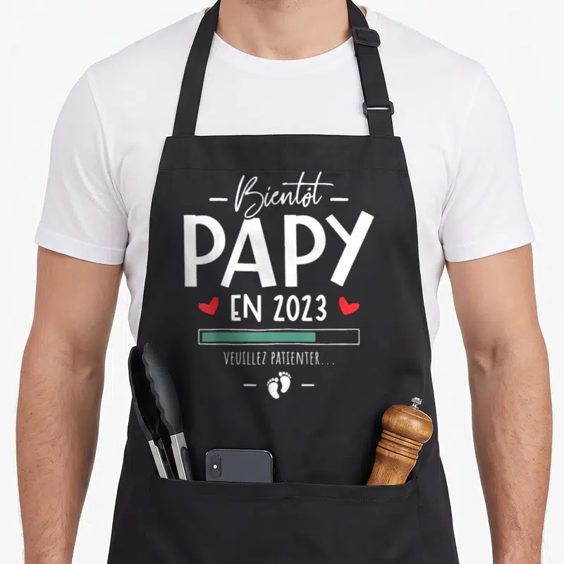 Tablier de cuisine noir avec texte blanc Bientôt Papy en 2023 et icônes de petits pieds rouges