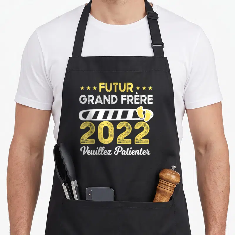 Tablier de cuisine noir avec l'inscription Futur Grand Frère 2022 Veuillez Patienter et deux traces de pas jaunes sur un bandeau blanc.