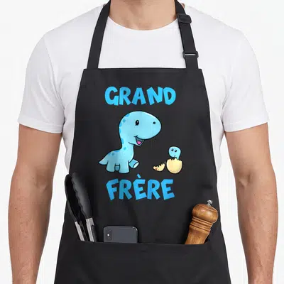 Tablier de cuisine noir avec un dinosaure bleu souriant et un œuf éclos portant l'inscription Grand Frère en lettres capitales.