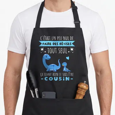 Tablier de cuisine noir avec deux dinosaures bleus et le texte « Ça tombe bien je vais être cousin ».